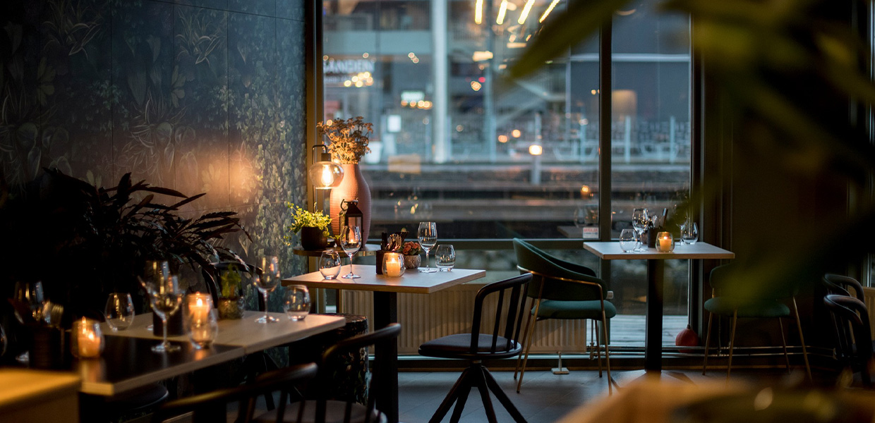 Fredelig spisestue med et koselig arrangement av bord foran en stor vindusvegg, innredet med blomster og stearinlys.