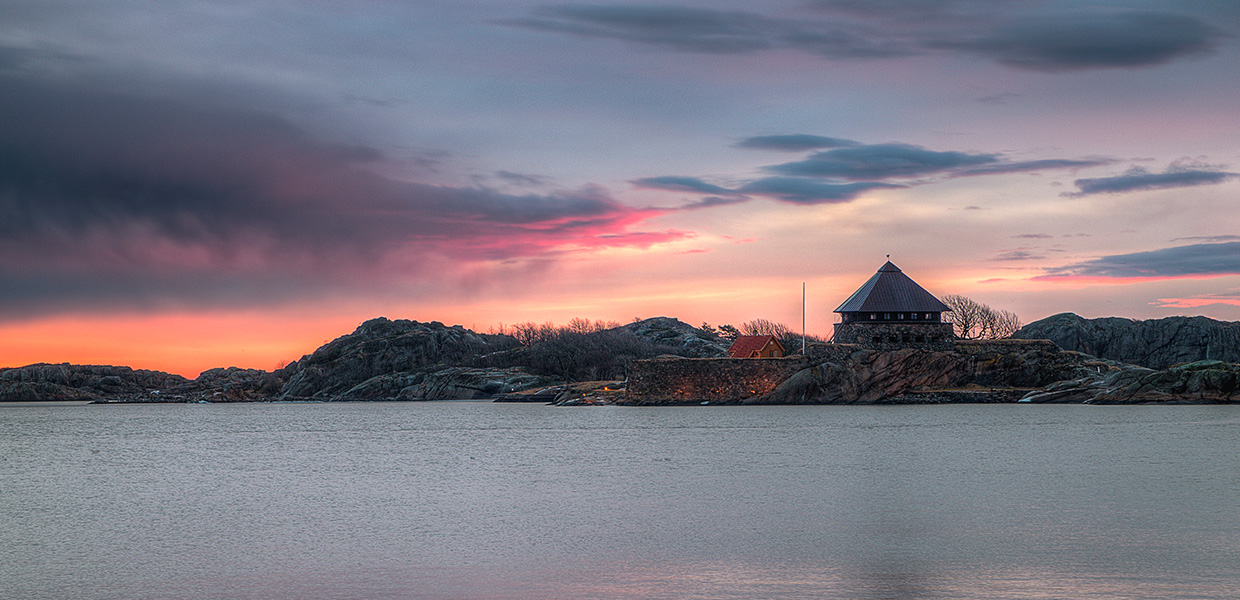 Vinterlandskap i Stavern ved solnedgang. Et rundt steintårn med konisk tak står på en odde omgitt av svaberg og bart fjell. Himmelen er dramatisk farget i rosa, oransje og blå toner, og det stille vannet reflekterer de varme fargene.