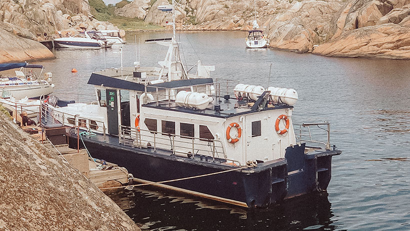 Et passasjerfartøy ligger ved kai i en havn omgitt av klipper. Skipet har sikkerhetsutstyr og oransje detaljer, klare for å transportere passasjerer som samles på brygga, og hvite båter som flyter ved siden av.