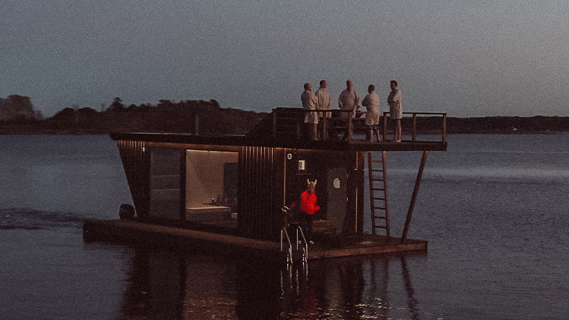En flytende badstue ved kysten omgitt av vann. Badstuen har et moderne design med en terrasse på toppen hvor personer oppholder seg. Vannsnivået reflekterer kveldslys.