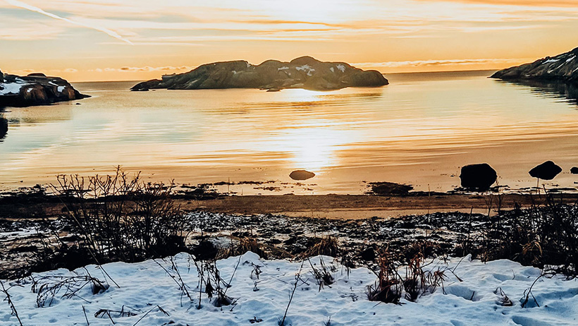 strand med snø på og utsikt mot holme i kveldssol